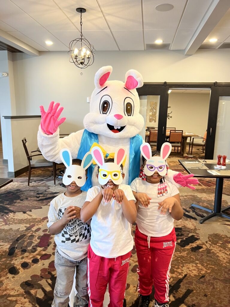 Three children wearing bunny masks pose with a person in a large bunny costume. The group stands indoors on a patterned carpet, and the children are making bunny ear gestures with their hands.