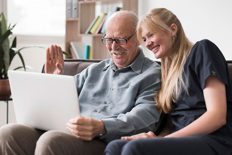 An elderly man and a young woman sit together on a couch, smiling at a laptop screen. The man waves at the screen, suggesting they are on a video call. Bookshelves and a plant are in the background.