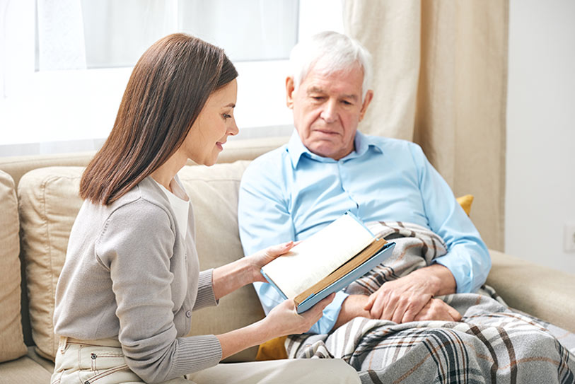 A young woman sits on a couch next to an older man covered with a blanket, showing him an open book and smiling as he listens attentively. They appear to be in a cozy, well-lit living room.