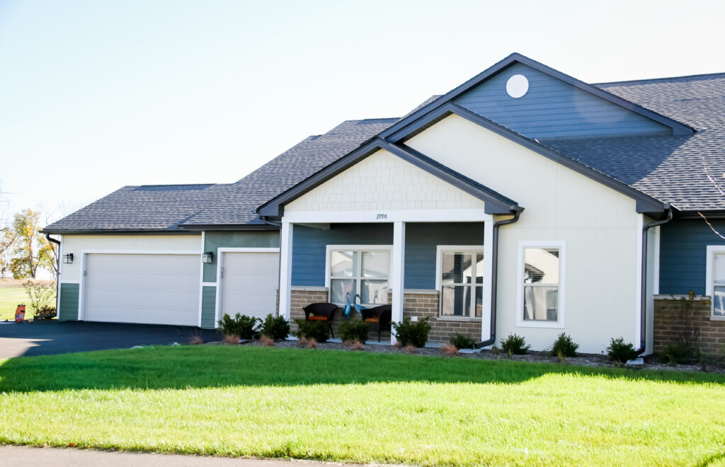 A modern single-story house with blue and white siding, a covered front porch, green lawn, shrubs along the front, and a large three-car garage on the left side.